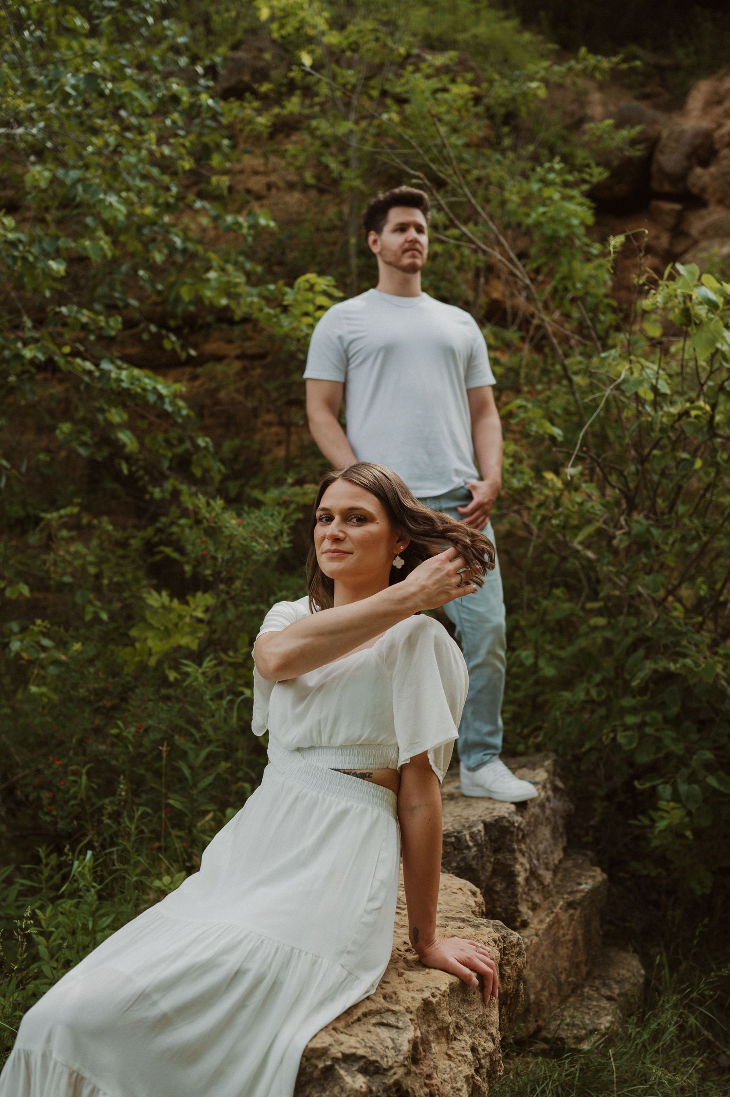 A couple poses on rocky terrain with one seated and one standing among lush greenery.