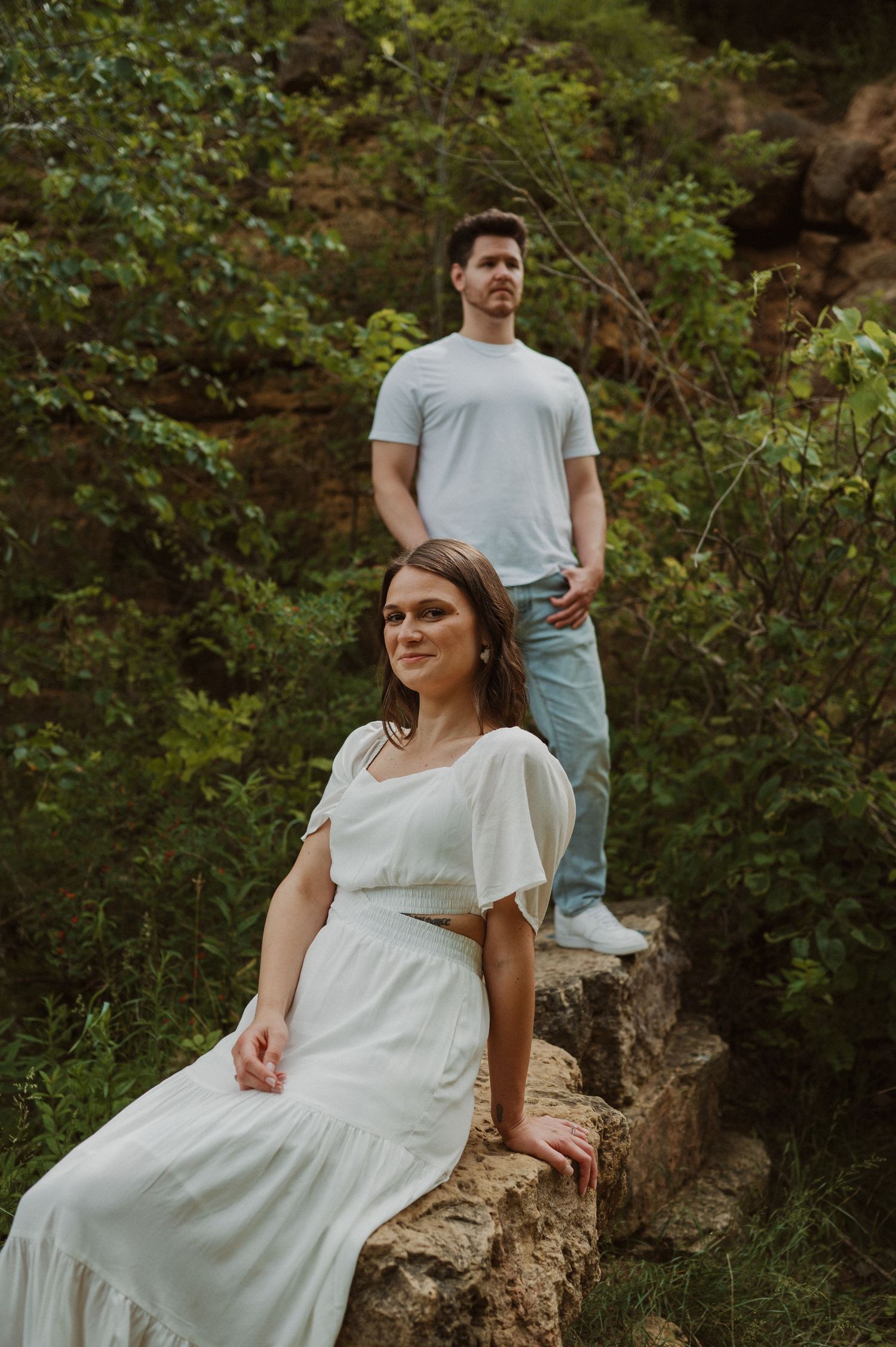 Two people pose on rocky terrain with one seated and one standing against natural scenery.