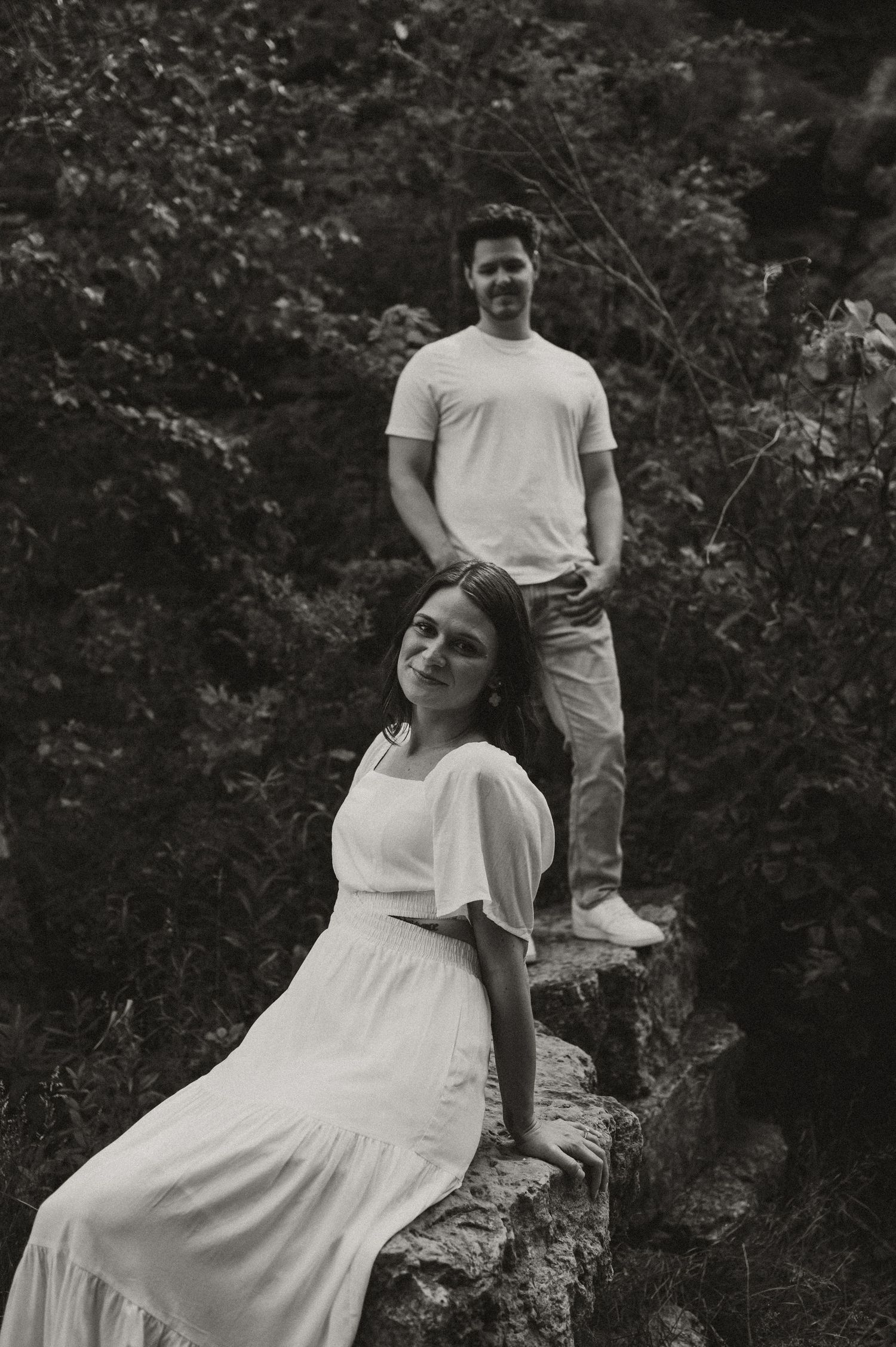 A couple poses in a wooded area for casual outdoor photos, with one person sitting on a stone ledge.
