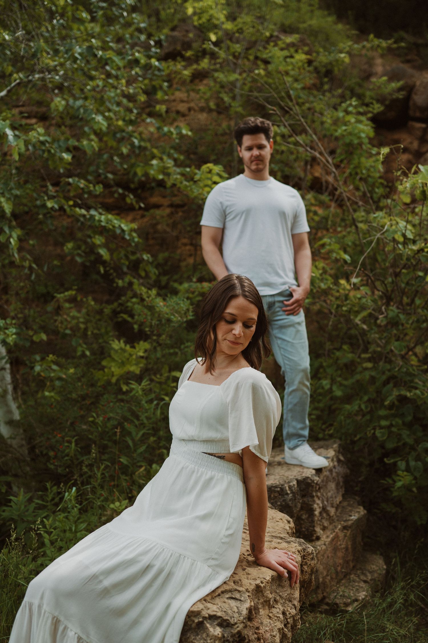 A person in a white dress sits on a rocky ledge while someone stands behind in a natural setting.