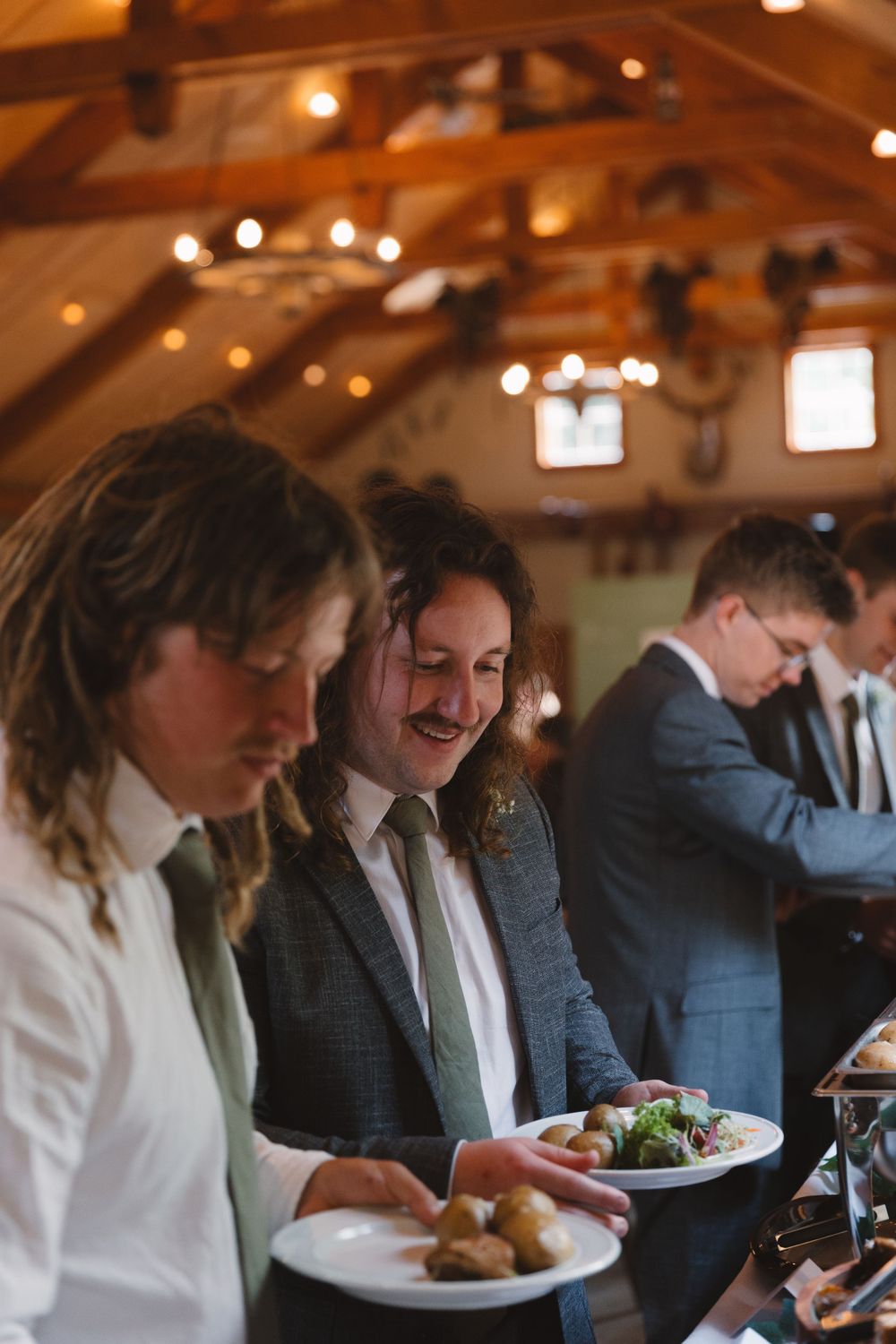Wedding guests serving themselves food in rustic venue with exposed wooden beams.