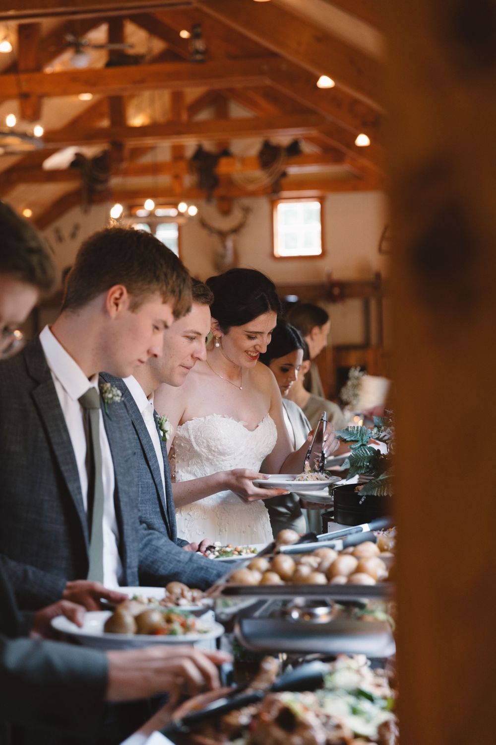 Wedding guests selecting food from buffet line under rustic wooden beams.