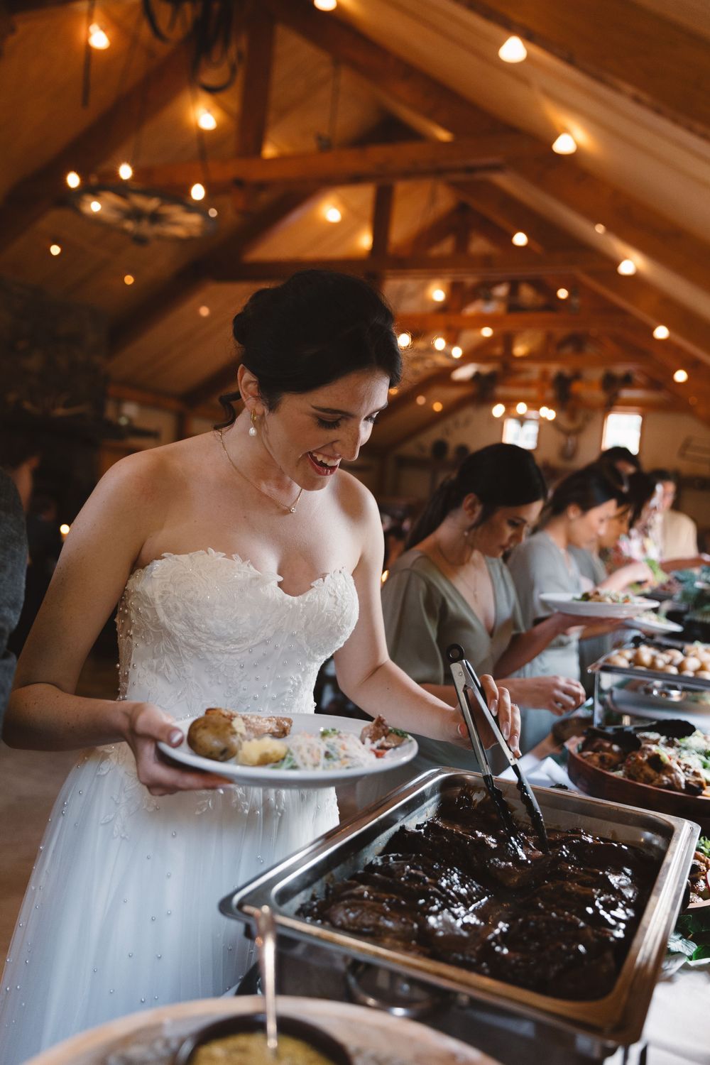 A bride in a white strapless gown serves herself food from a buffet line under rustic wooden beams with twinkling lights.