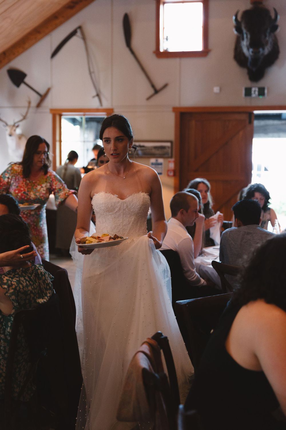 Bride in white dress walking through rustic dining room with mounted antlers and farm tools on wall.