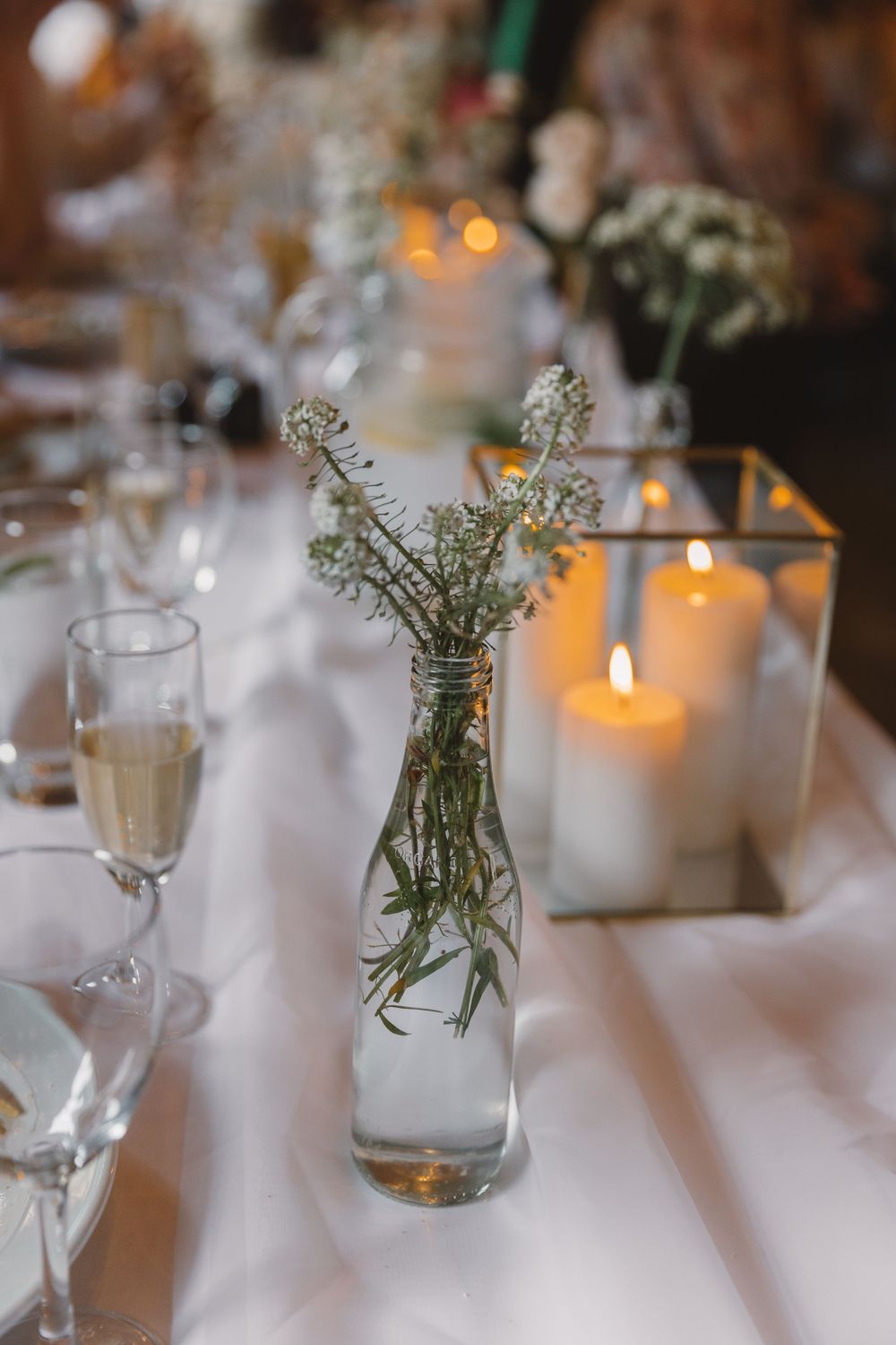 Elegant table setting with white candles in glass holders and delicate flowers in clear vases.