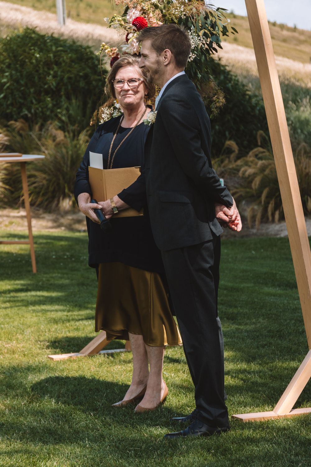 Wedding officiant and participant stand beneath wooden arch during outdoor ceremony.