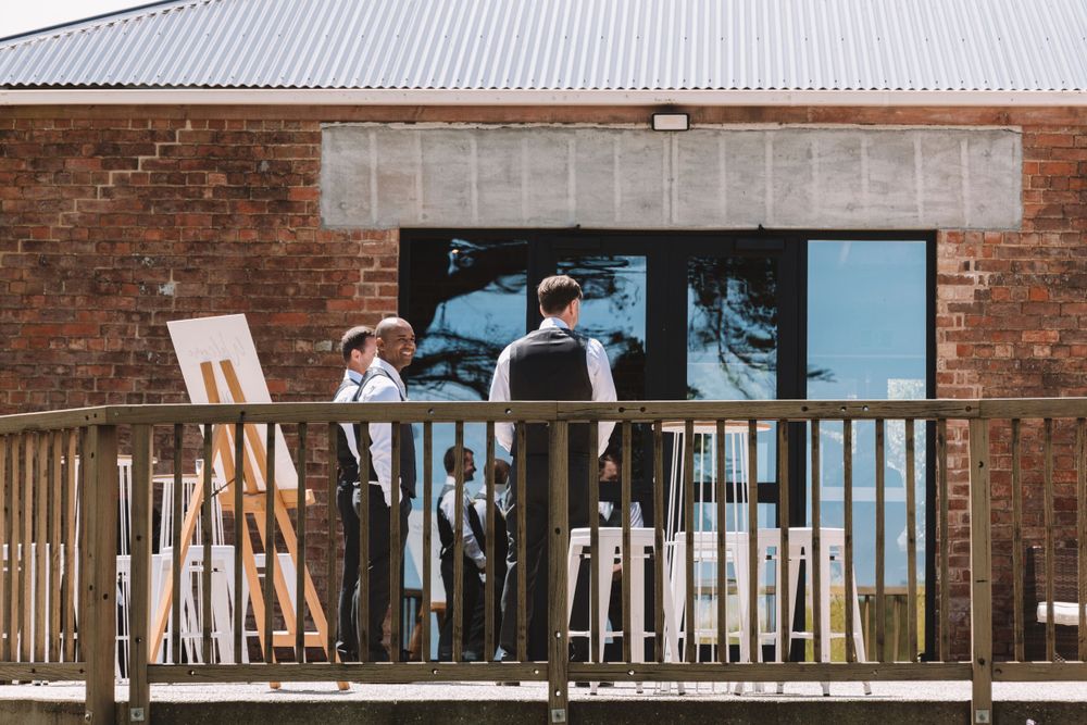 Wedding attendees stand on wooden deck of brick building during daytime celebration.