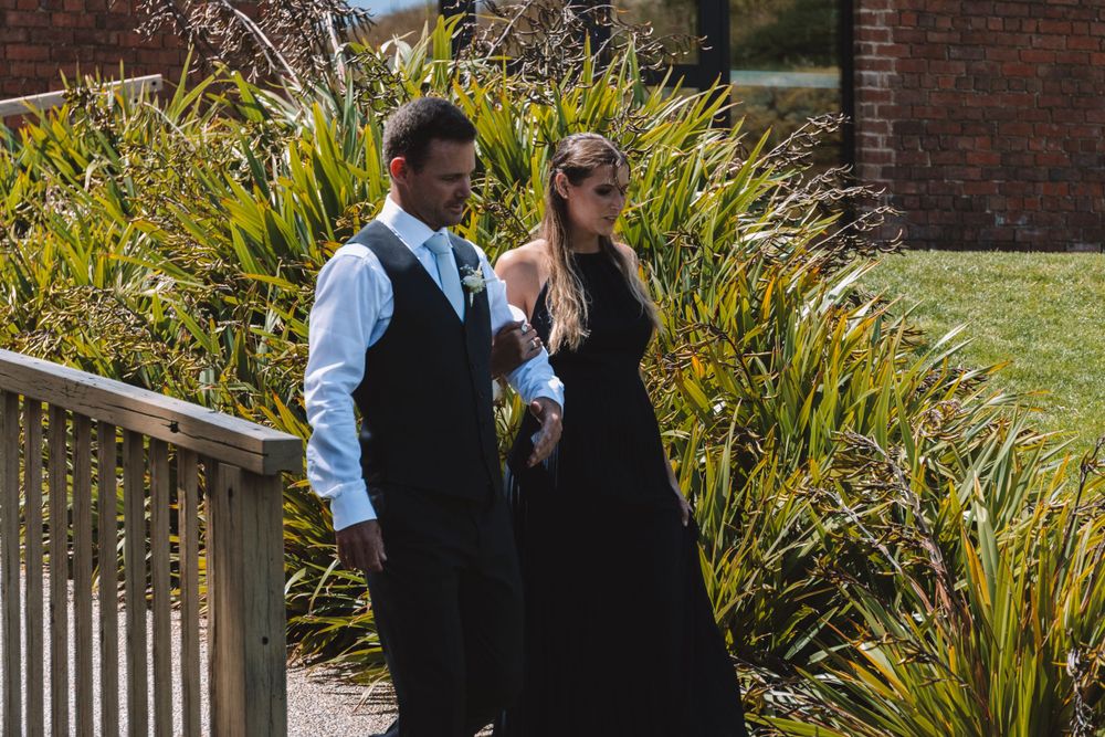 Formal wedding attendants walk along wooden deck with blooming plants in background.