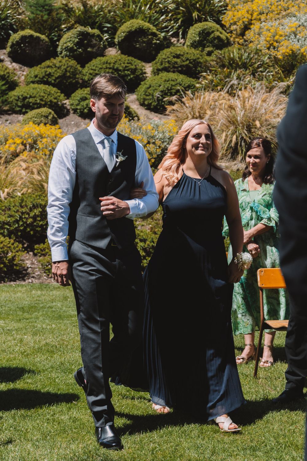 Wedding party members in formal attire walk through manicured garden during sunny celebration.