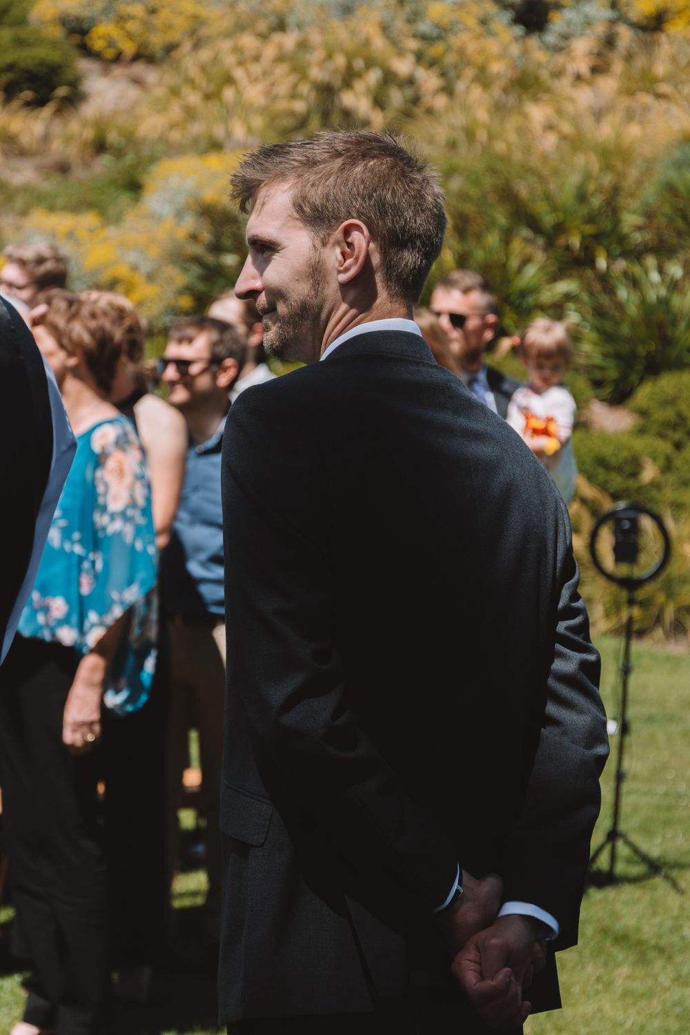 Wedding guest in dark suit stands among ceremony attendees in outdoor garden setting.