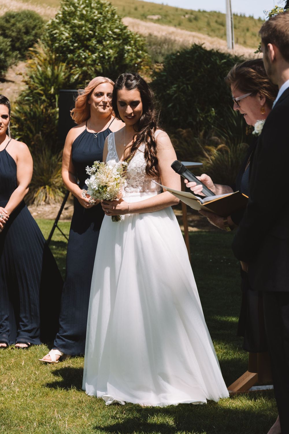Wedding ceremony in progress with attendants and white-gowned bride holding delicate bouquet.