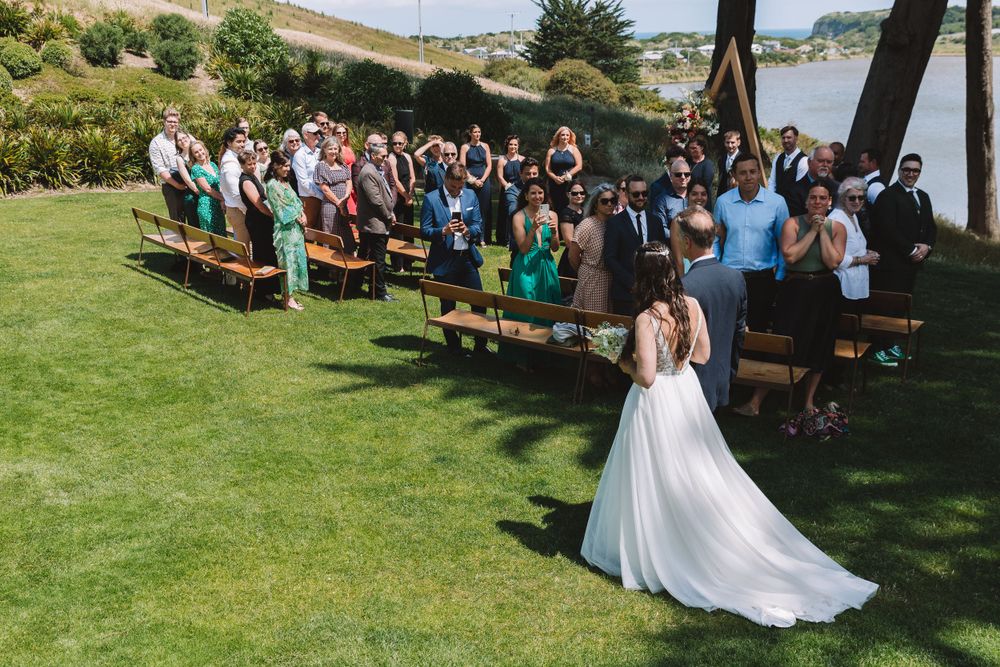 Wedding guests seated on wooden benches during an outdoor ceremony with a flowing white dress in view.