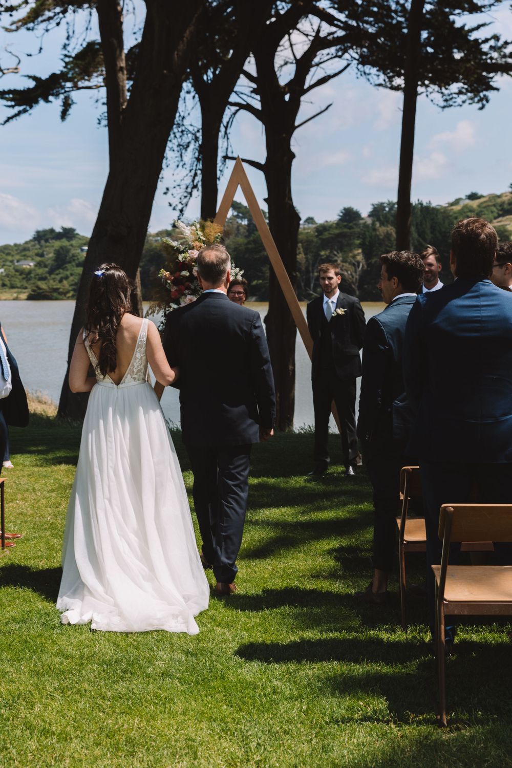 An outdoor wedding ceremony taking place under tall trees beside a lake.