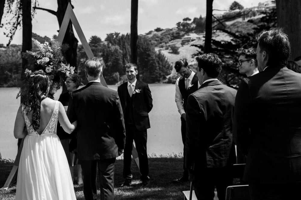 A black and white photo of a wedding ceremony gathering under trees near water.
