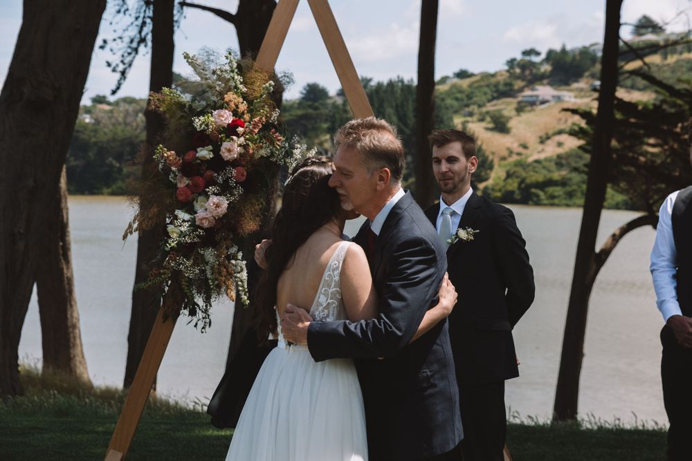 An intimate wedding ceremony moment under a wooden arch with a lake backdrop.