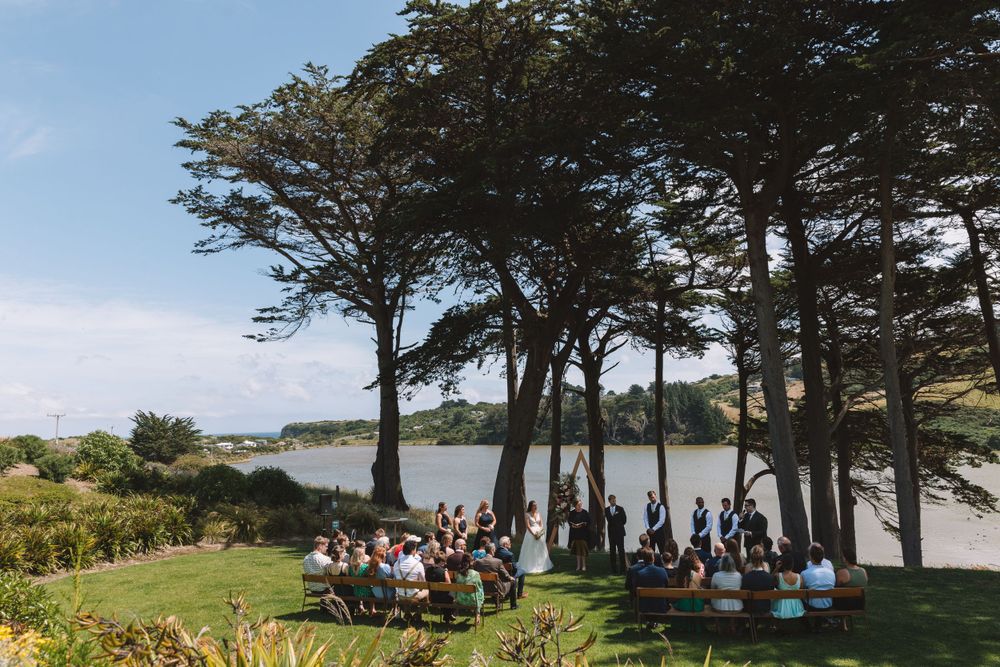 Wedding guests gathered for an outdoor ceremony with a scenic water view through tall trees.