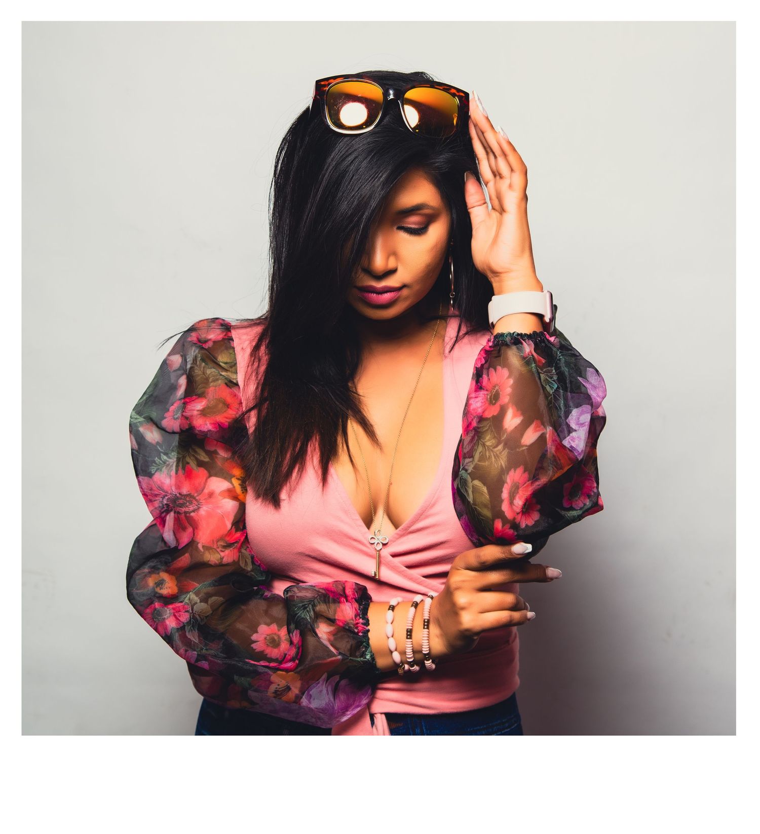 A model wearing a pink top with a floral sheer blouse, sunglasses on head and jewelry accessories against a neutral background.