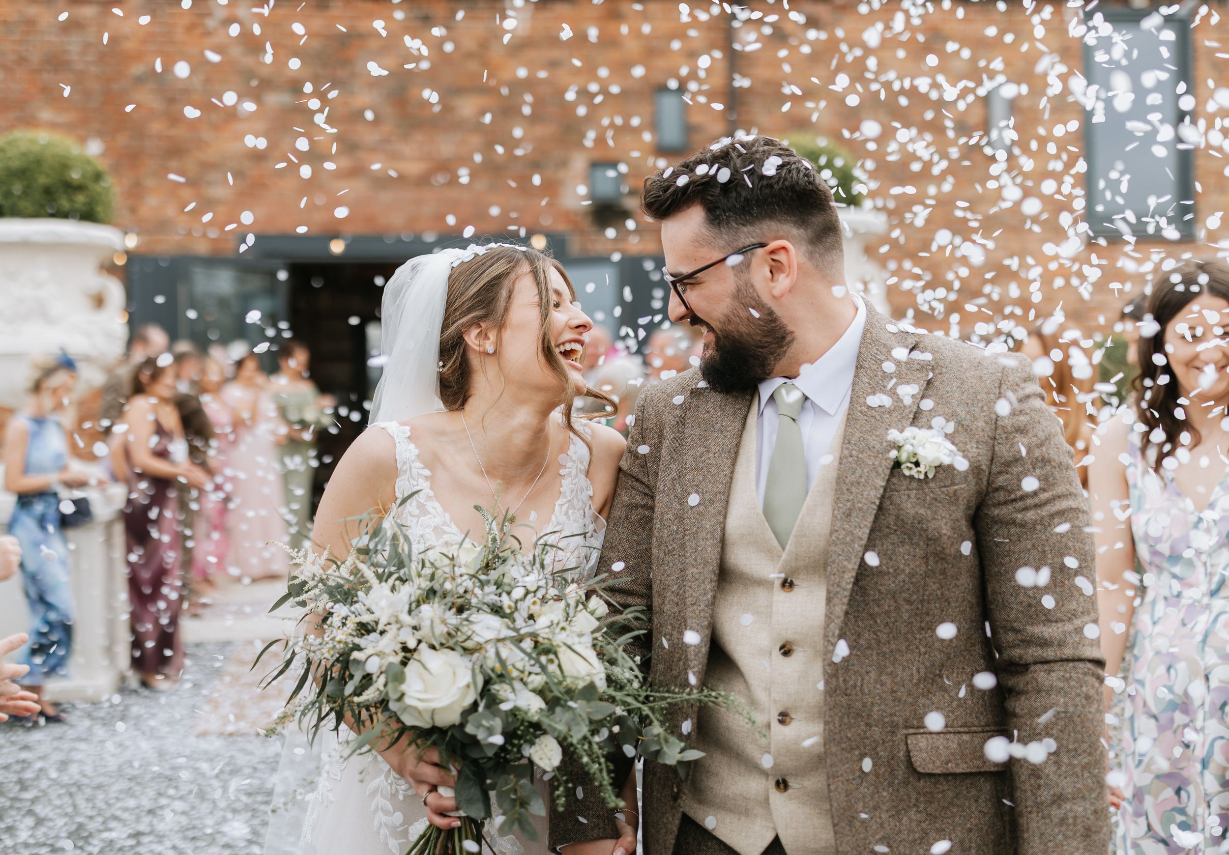Couple shares a joyful moment as wedding guests shower them with white confetti outside a brick venue. The venue is Burlton Manor.
