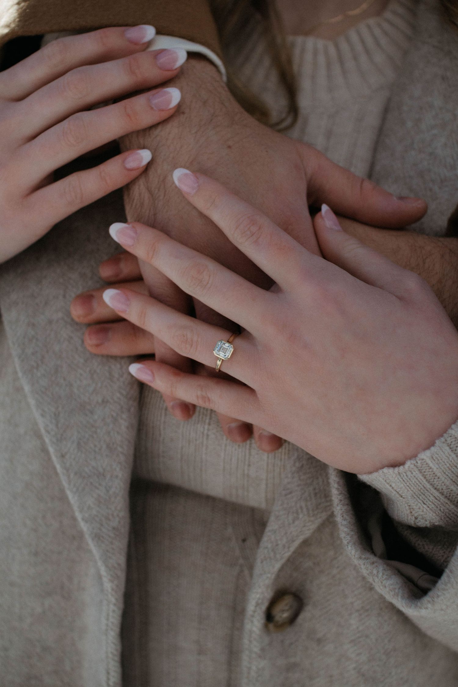 Close-up of hands with white manicured nails and engagement ring resting on a beige sweater background. engagement