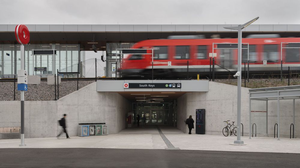 Modern train station entrance with red commuter train passing above and pedestrians using an underground passage.