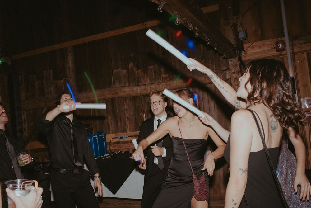 Party guests in formal attire wave glowing LED foam sticks while dancing in rustic wooden venue.
