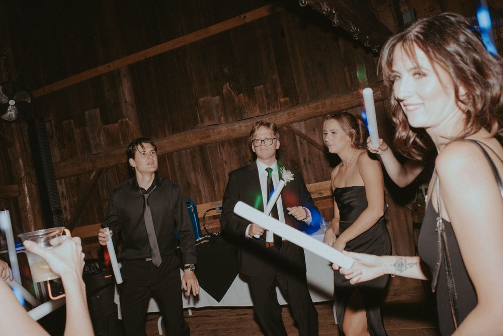Guests dance while holding light-up foam sticks at evening celebration in rustic venue.