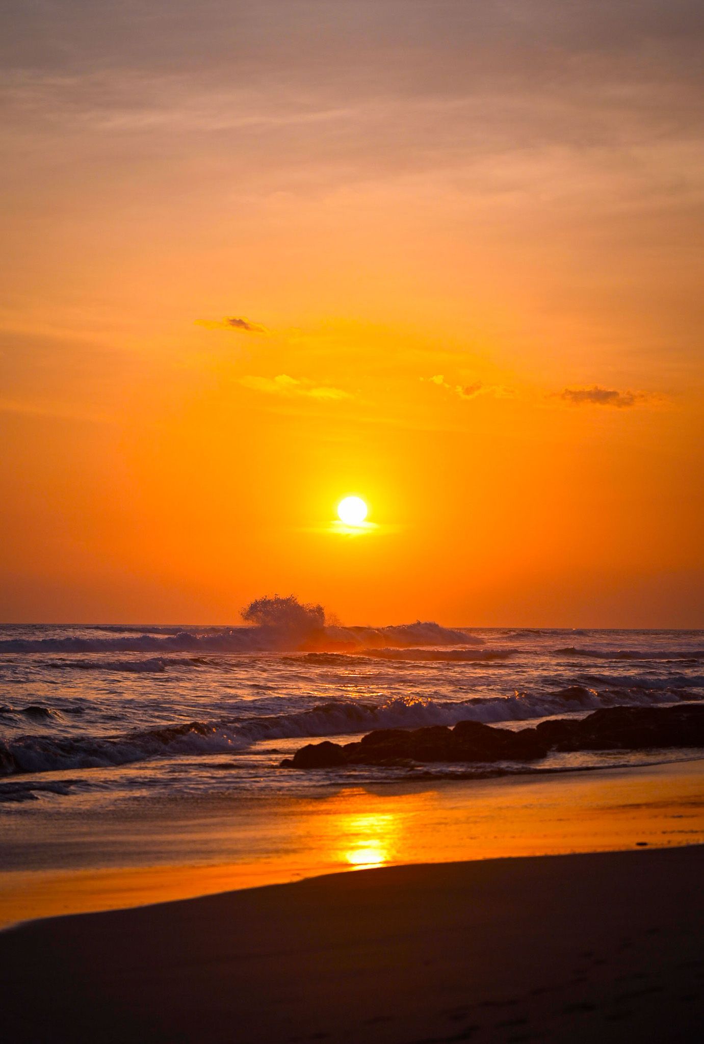 A dramatic orange sunset over the ocean with waves crashing against rocks and reflecting golden light on the beach.