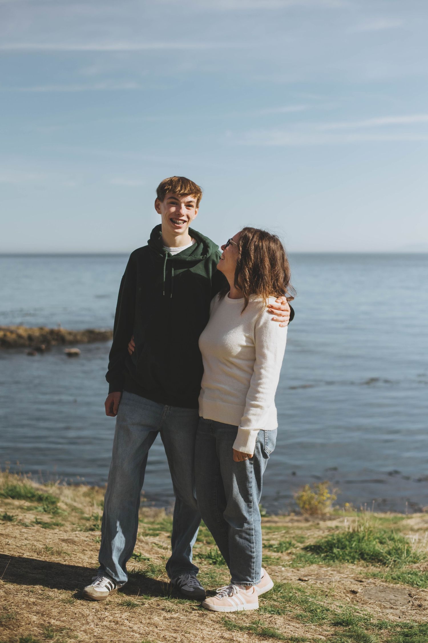 A mother and her teenage son share a lighthearted moment, smiling and laughing together as they stand arm in arm.