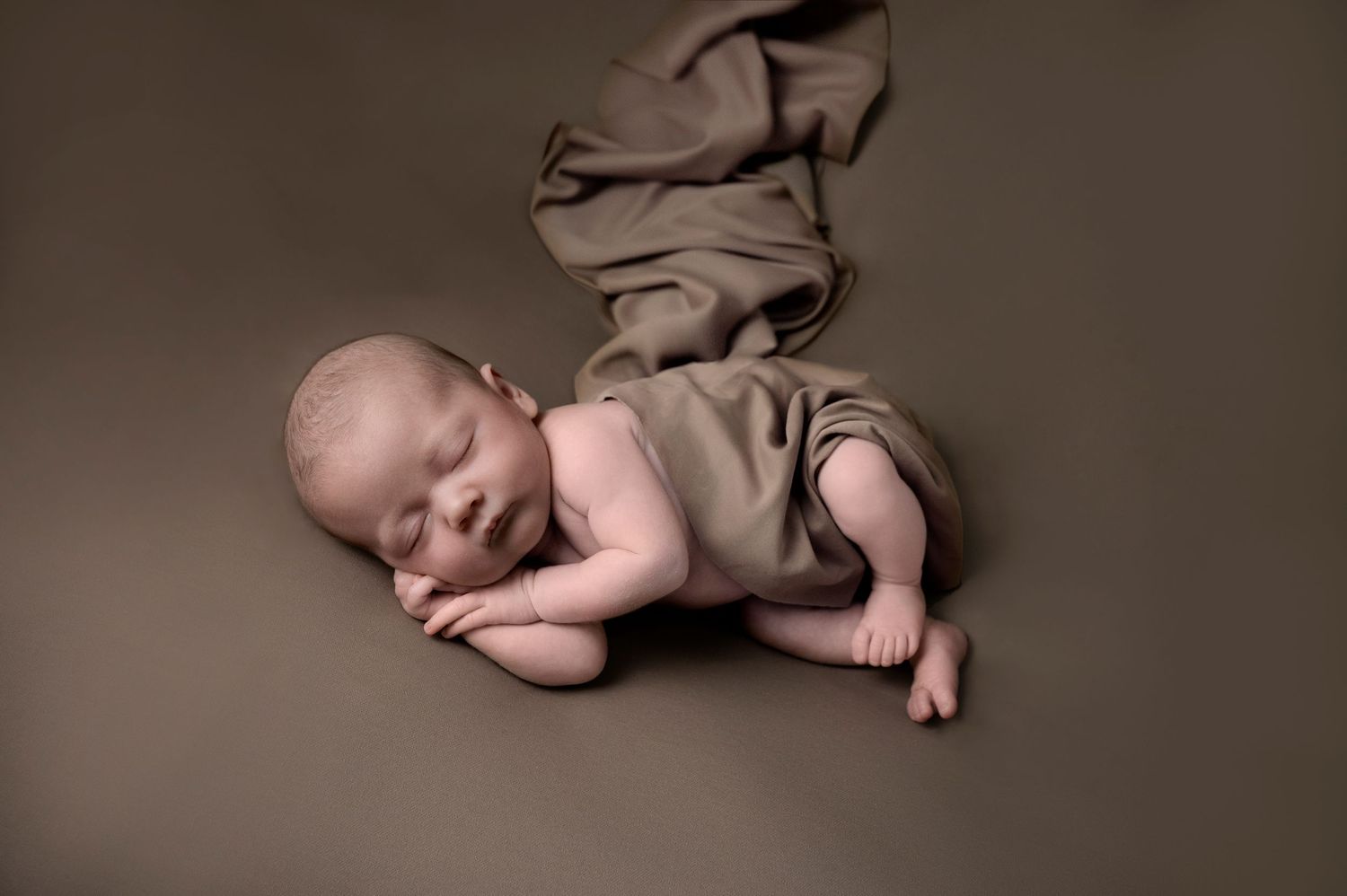 Newborn baby sleeping peacefully in brown wrap on dark brown backdrop.