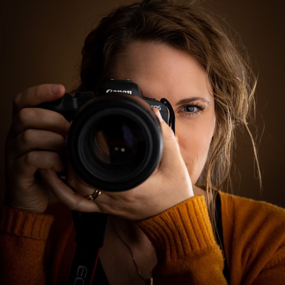 Close-up portrait shot of someone holding a professional camera while wearing an orange sweater.