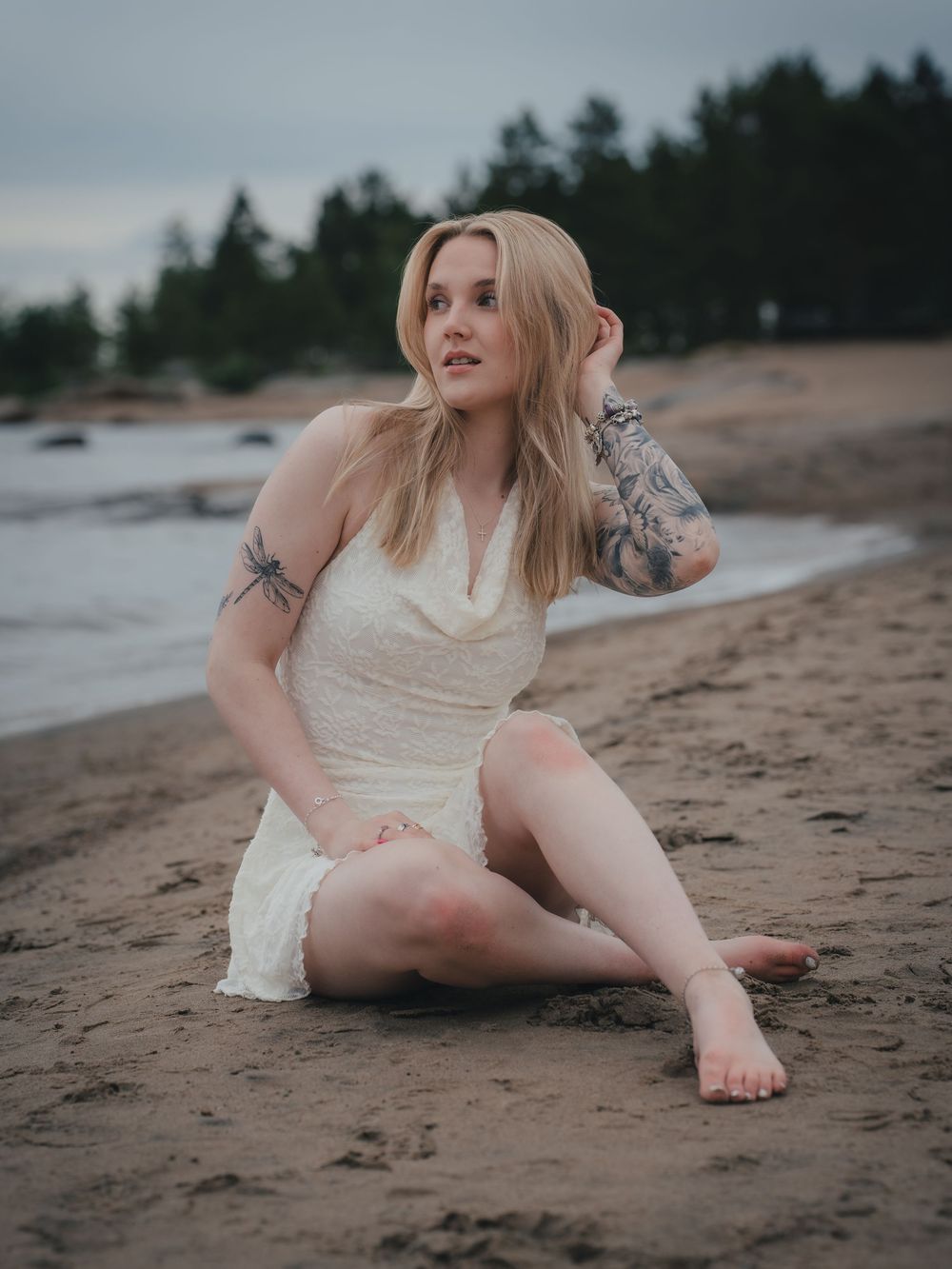 A person in a white dress sits on a sandy beach at dusk with pine trees and water in the background.