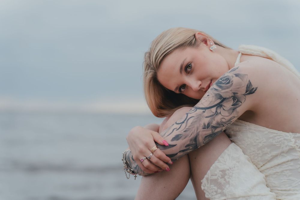 A person with arm tattoos poses thoughtfully near the ocean in soft natural lighting at sunset.