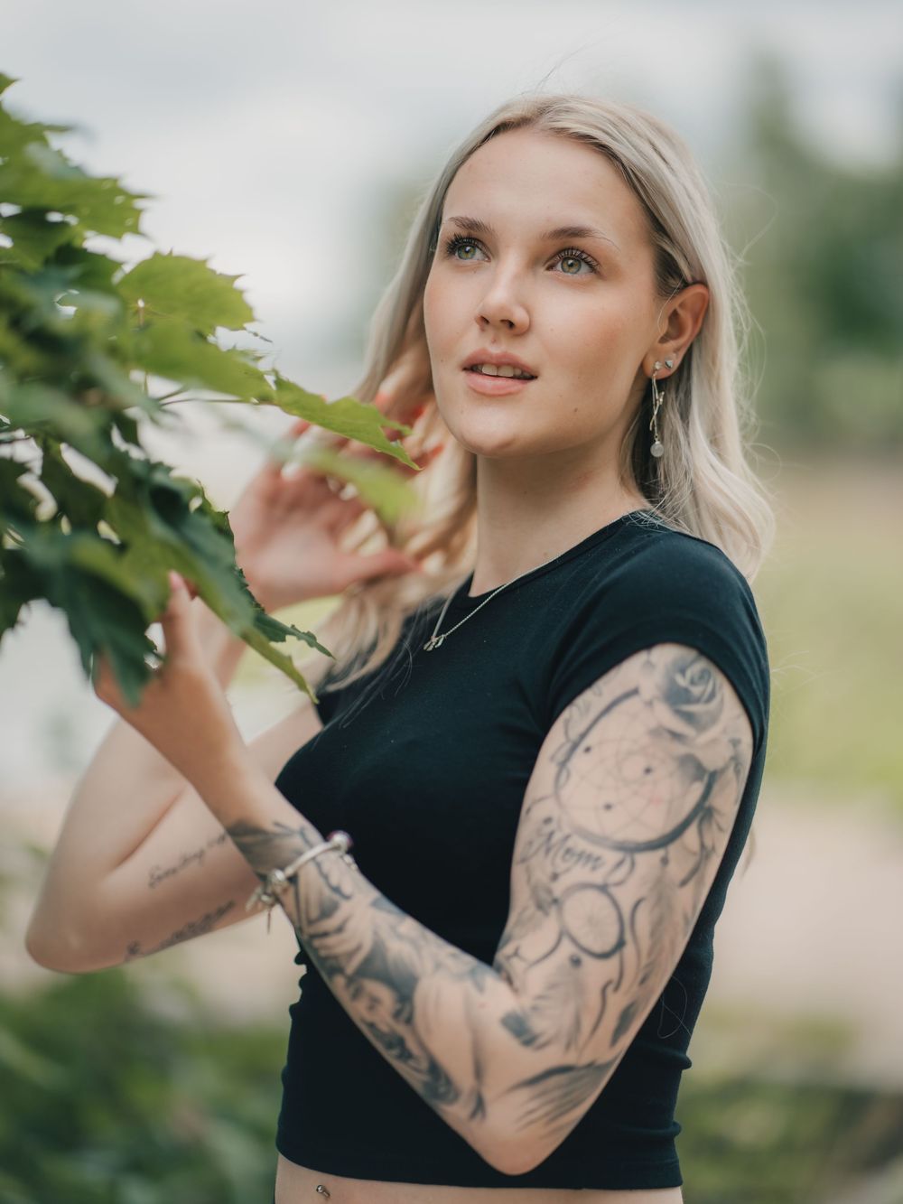 Blonde woman in black top with intricate sleeve tattoo interacts with green foliage outdoors in soft natural lighting.