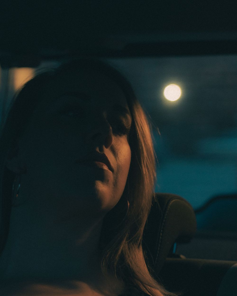 Silhouette at dusk with bright full moon glowing in a dark teal sky through car window.