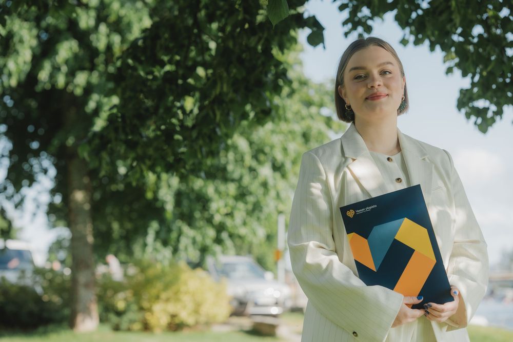 Professional in white blazer holding folders standing outdoors under green trees on sunny day.