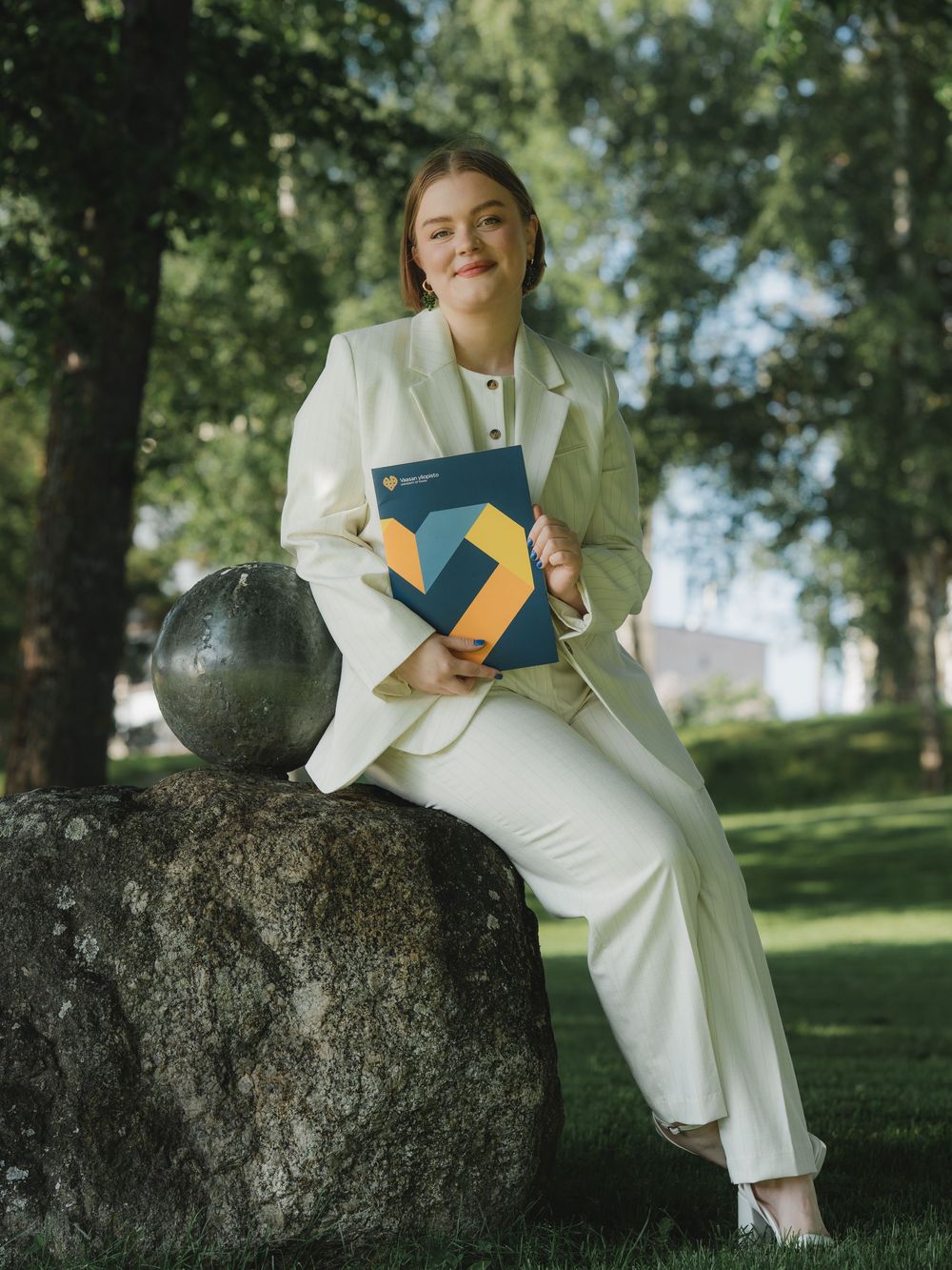 Person in an all-white outfit sits on a stone sculpture while holding a branded folder in a park setting.