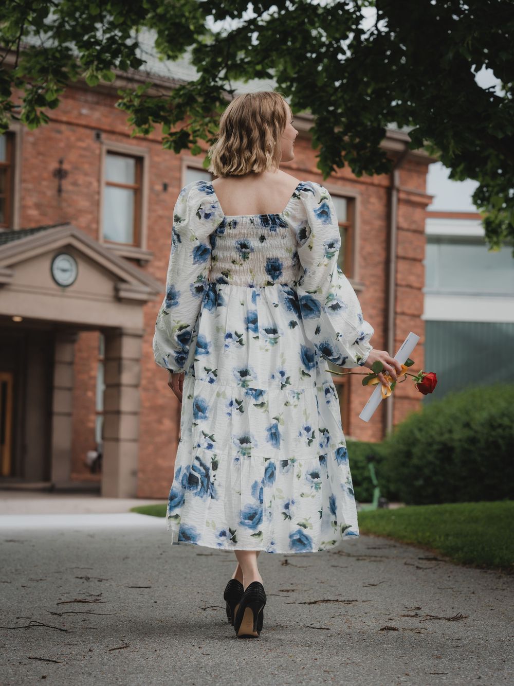 White and blue floral dress flows gracefully while walking along a brick building pathway in elegant fashion.