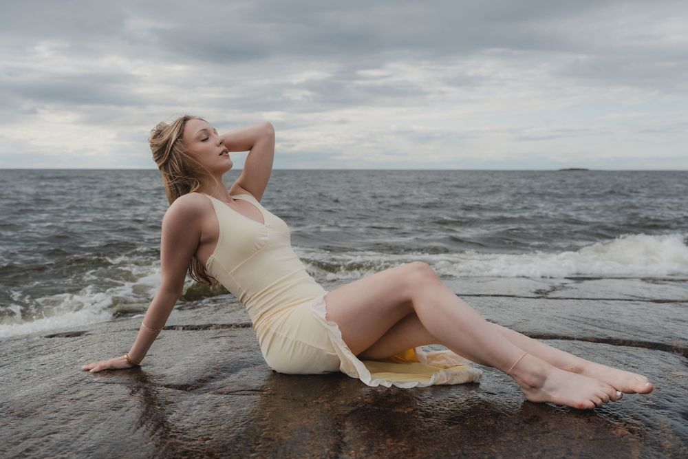 A person in a cream dress reclines on dark coastal rocks as waves crash against the shoreline under cloudy skies.