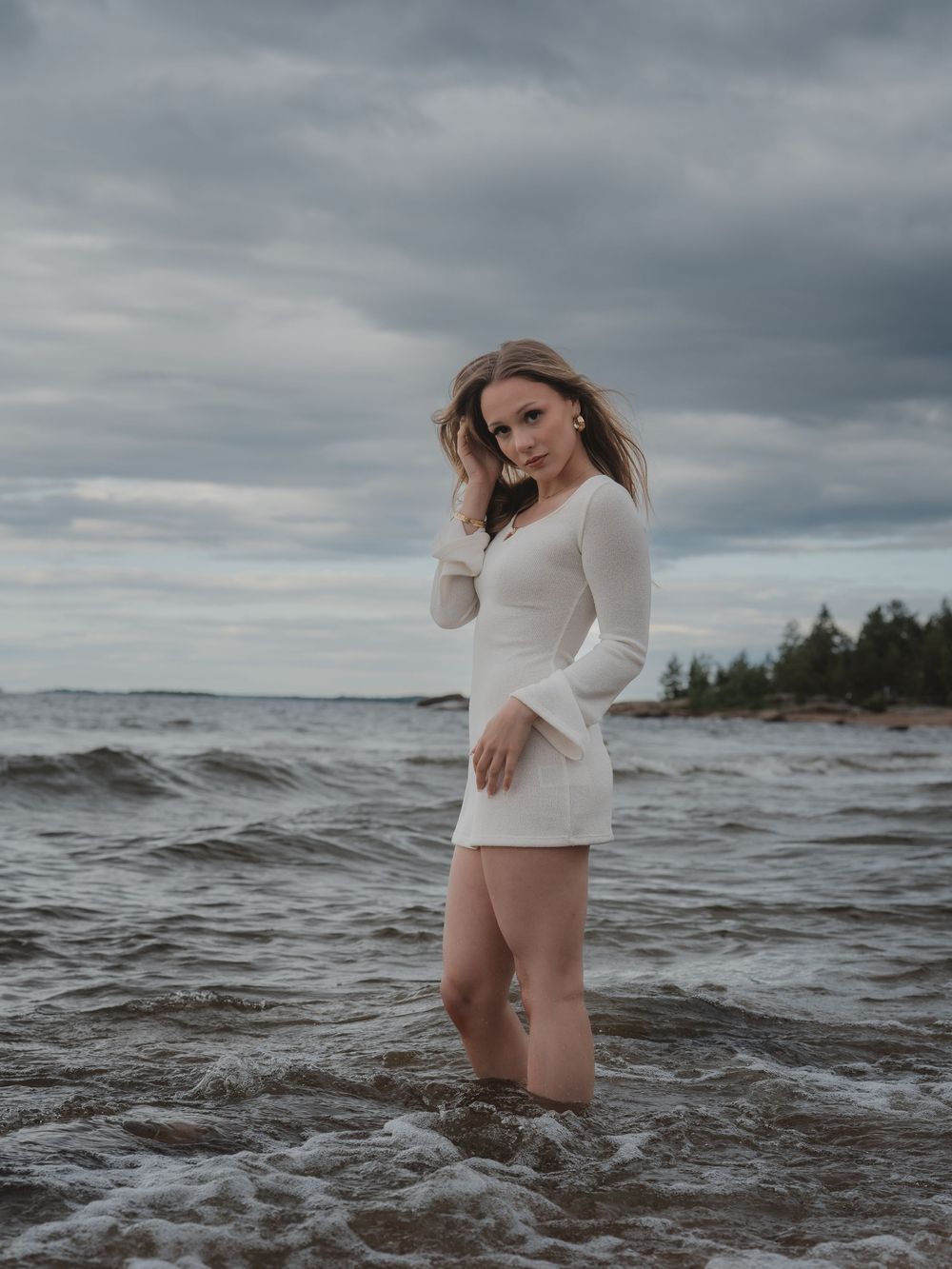 Figure in white fitted dress stands in moody ocean waters against stormy sky and pine-lined shore.