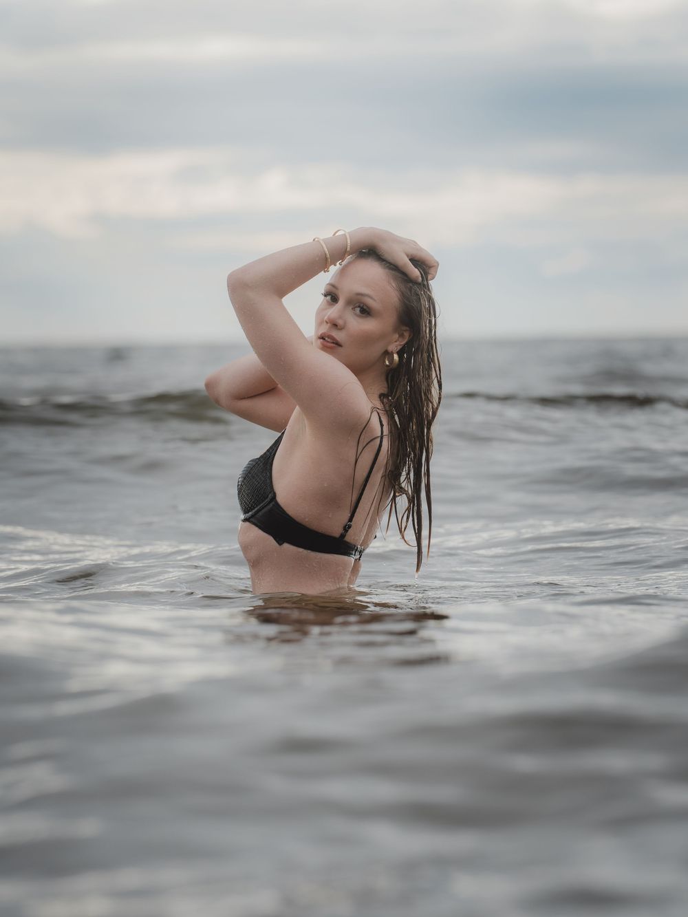 A person emerges from dark ocean waters in a black swimsuit during a moody overcast photoshoot session.