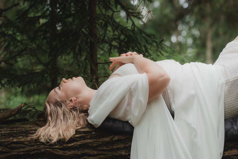 A bride in a white dress lies dreamily across moss-covered ground in a tranquil forest setting.