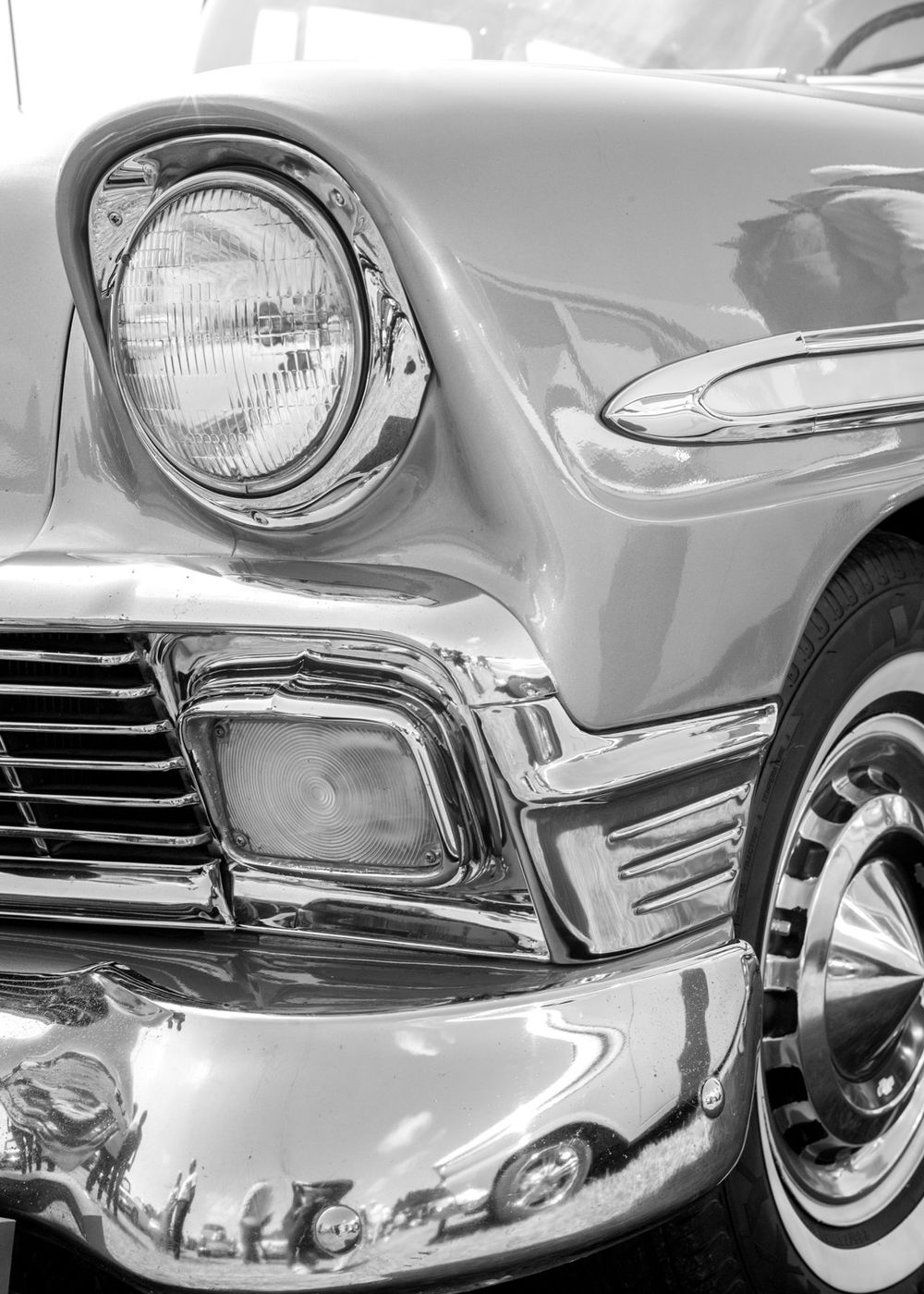 Close-up black and white photo of classic 1950s car headlight and chrome grille details showing vintage automotive design.