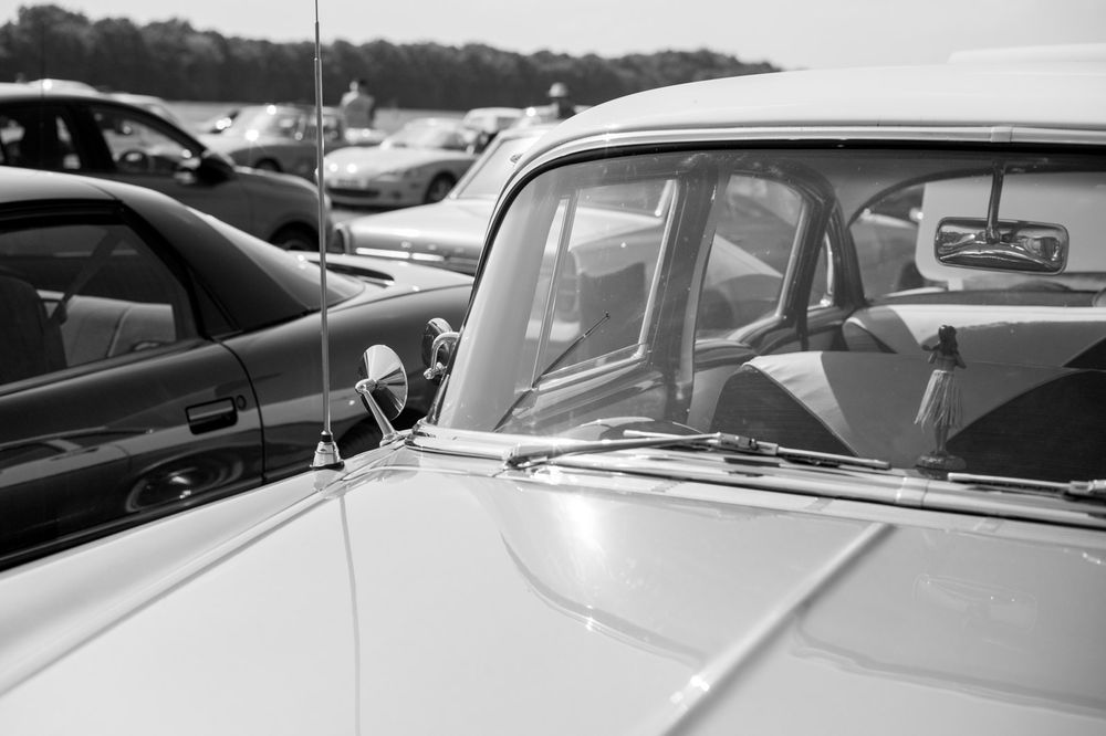 Black and white close-up detail of a vintage car's windshield and hood at an outdoor parking area.