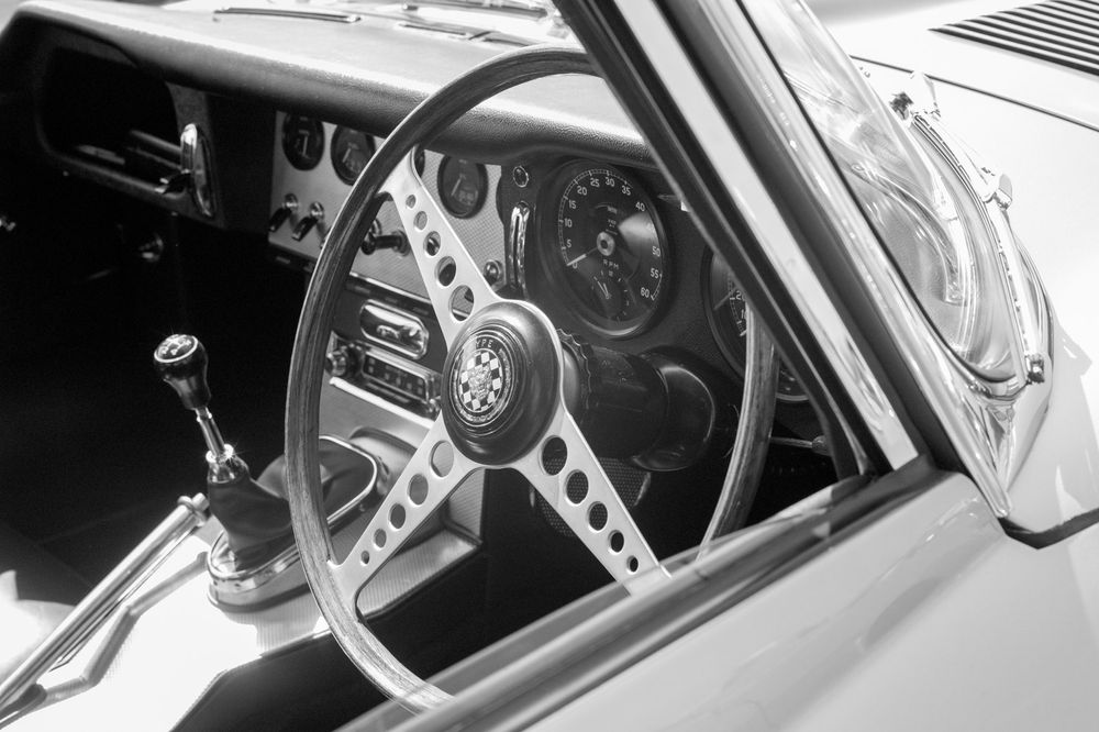 Interior view through car window showing vintage sports car steering wheel and dashboard in black and white photography.