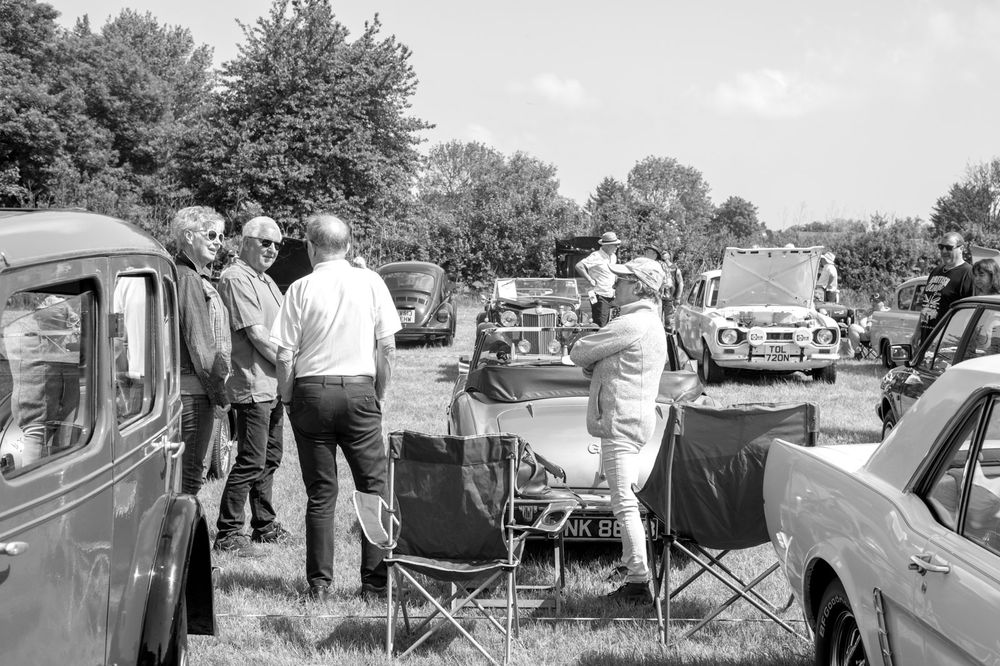 Vintage cars and people gathered at an outdoor event with folding chairs visible in black and white photography.