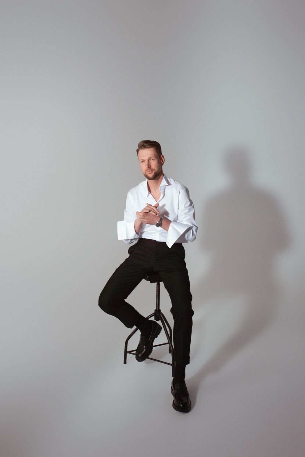Fashion studio photo of a person in a white dress shirt and black pants sitting on a stool against a gray background.