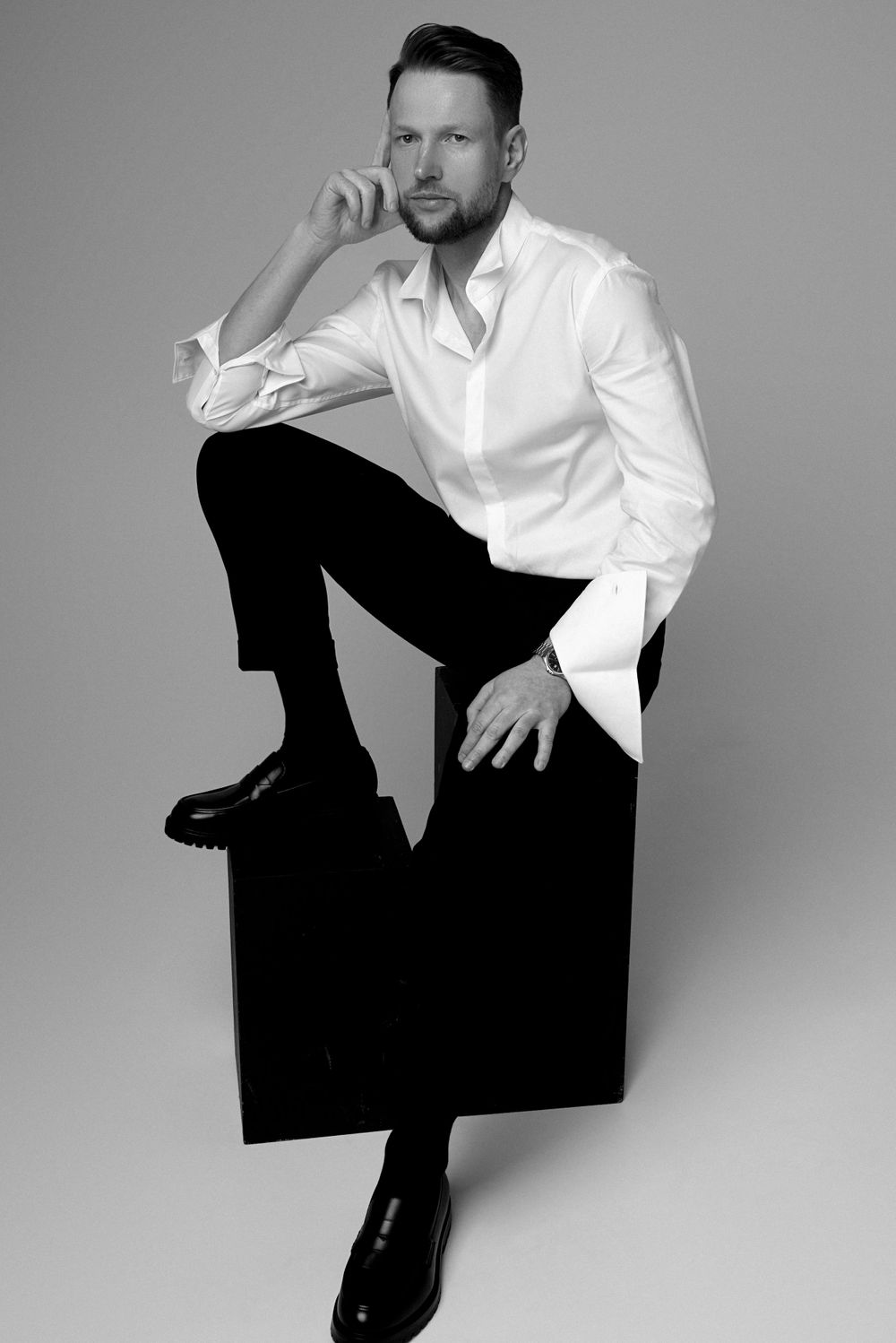 Black and white studio portrait of someone in a white button-down shirt and dark pants sitting on a dark cube.