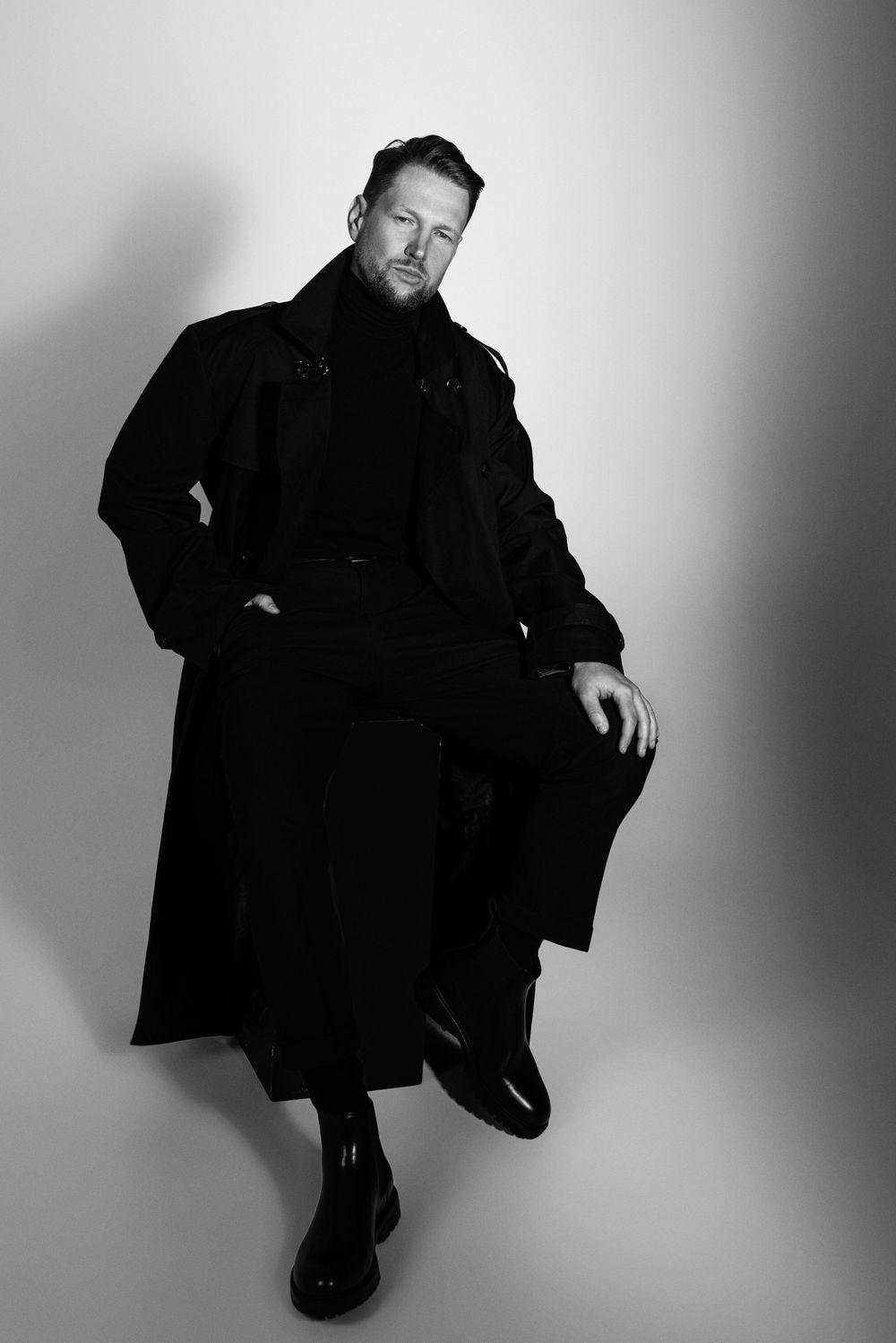 Black and white portrait of a person wearing a long dark coat while sitting in a relaxed pose against a gradient background.
