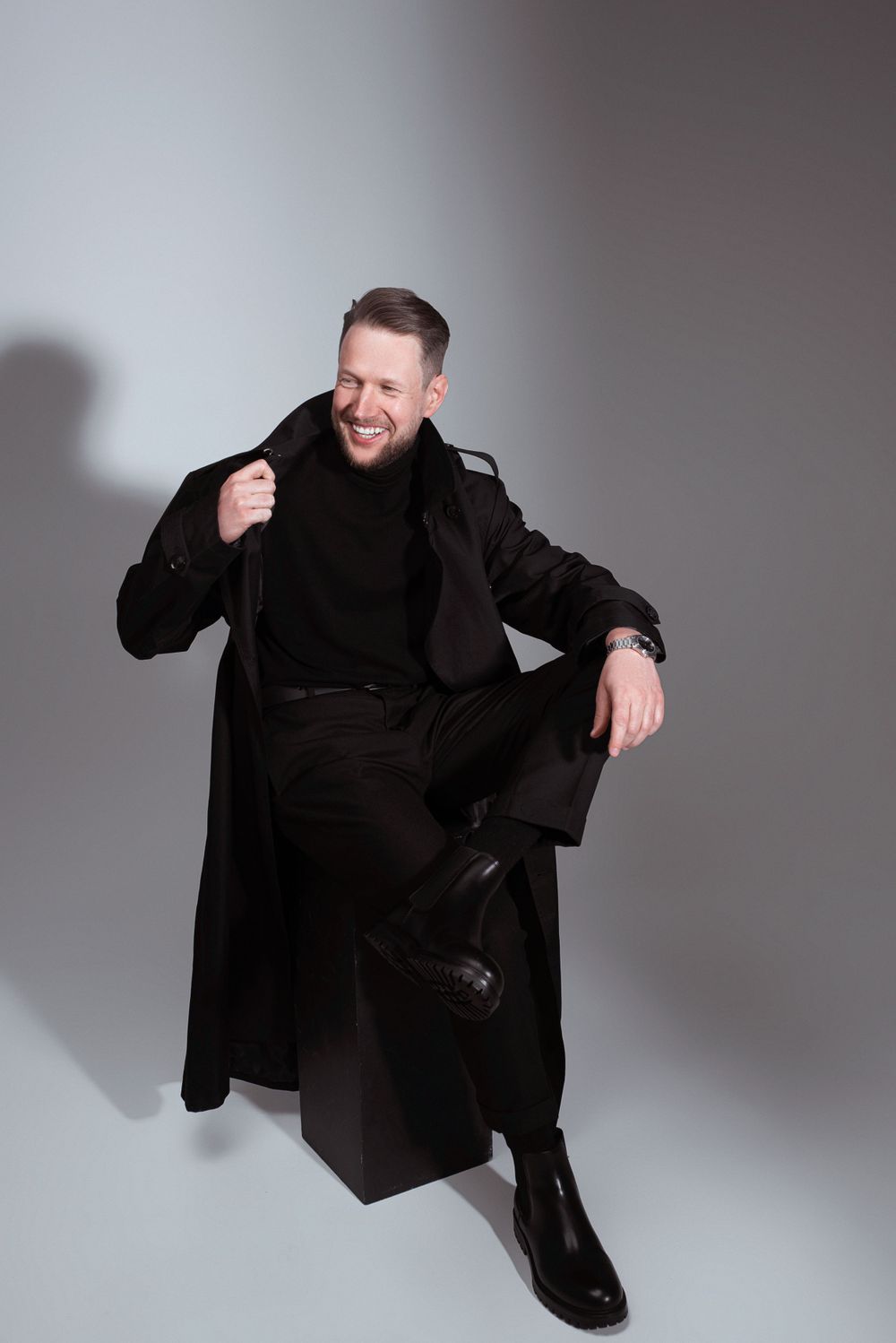 A person wearing a black winter coat and boots sits casually against a gray studio backdrop while smiling.