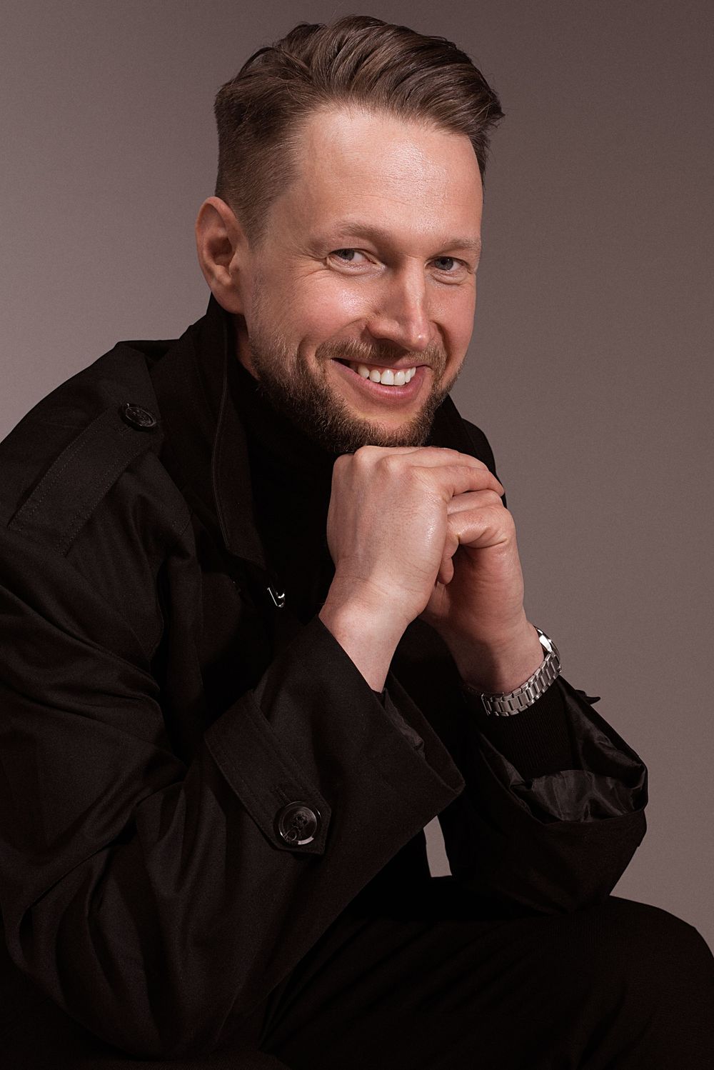 A person wearing a dark jacket poses for a professional portrait with hand on chin and warm smile against neutral background.